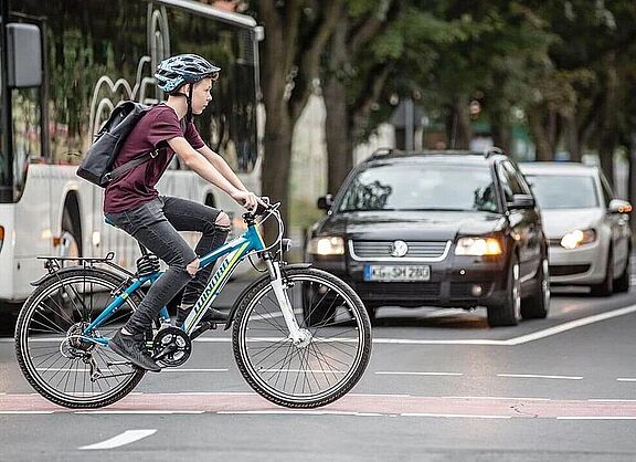 Junge fährt auf dem Fahrrad über einen rot markierten Radweg an wartenden Autos vorbei.