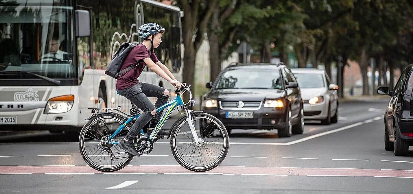 Junge fährt auf dem Fahrrad über einen rot markierten Radweg an wartenden Autos vorbei.