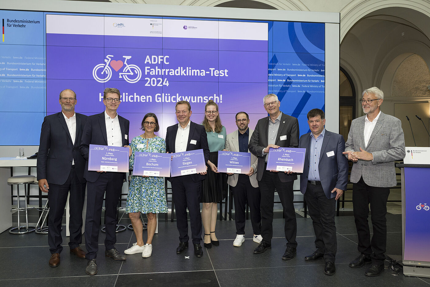 Neun Personen stehen in einer Reihe vor großer Leinwand mit "ADFC Fahrradklima-Test 2024 Herzlichen Glückwunsch!" und halten lila Urkunden, darunter Nürnberg, Bochum, Lahr, Witten und Rheinbach